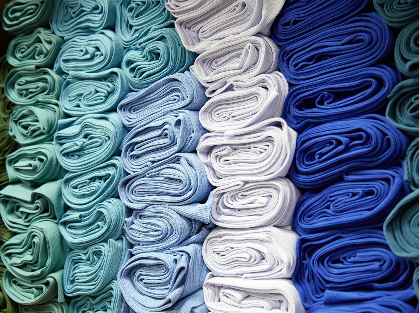 Rolos de tecido em tons de azul, verde-água e branco empilhados lado a lado